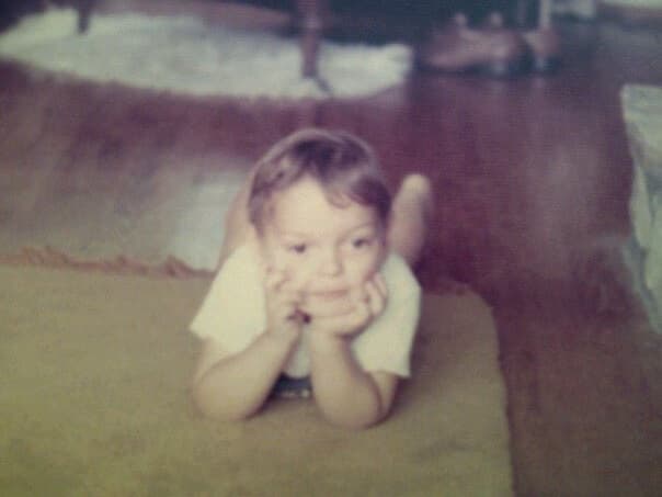 Andy as a toddler, chin in hands, lying on a rug in a 1970s living room