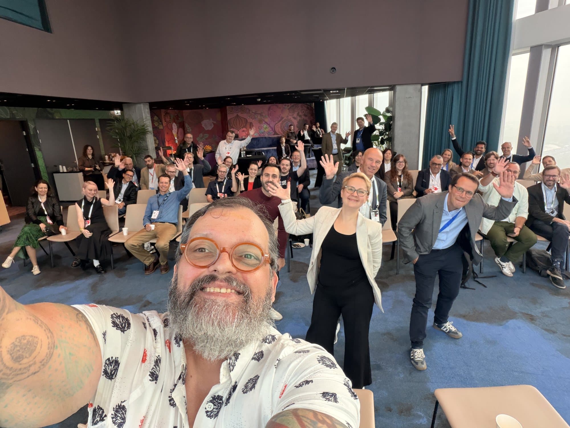 Andy Beach taking a selfie at the front of a conference room, the audience waving behind him