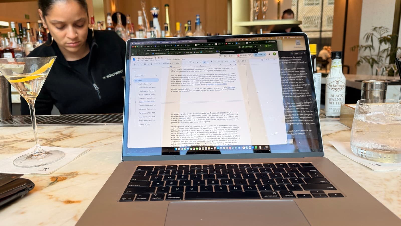 A laptop with an essay draft open on a marble bar top, a martini on one side and cocktail bitters on the other